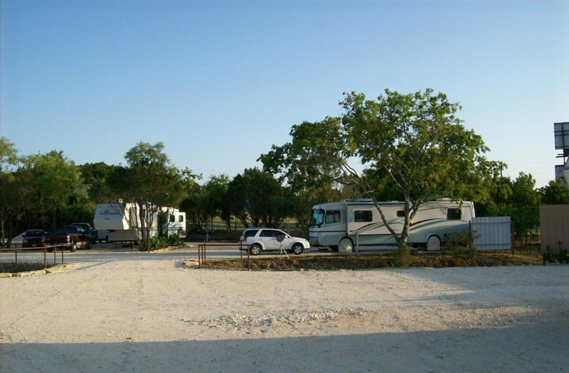 A motorhome and a fifth-wheel trailer parked among trees at the park, across a wide gravel drive